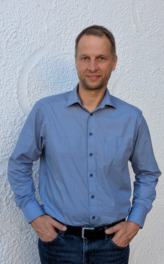 Bild Martin Köpernik Martin Köpernik mit blauem Hemd und Jeans, steht mit lächelndem Gesicht vor einer hellen Wand.
